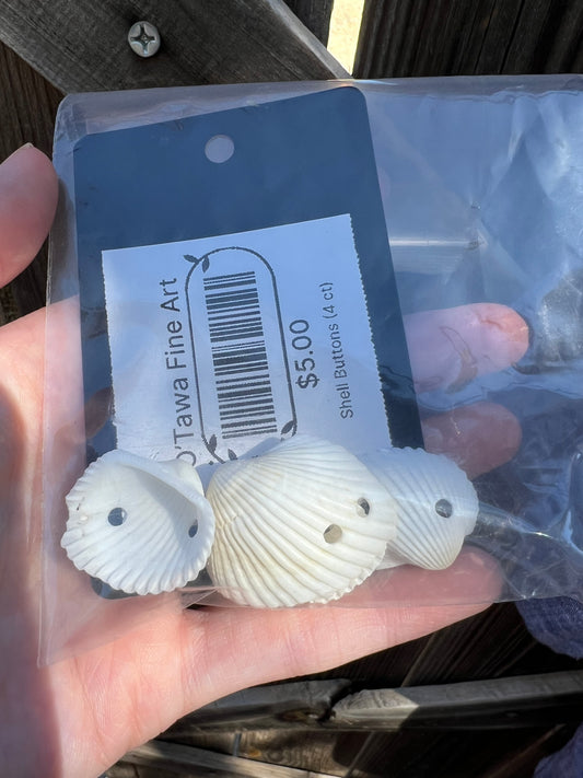A hand holding a clear plastic bag containing four small white shell-shaped buttons with pre-drilled holes, attached to a dark blue hang tag labeled O'Tawa Fine Art and $5.00.